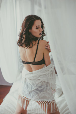 A young attractive girl with dark hair in black lingerie with evening make-up and jewelry and a white bathrobe on a white bed in the bedroom. beauty and fashionの写真素材