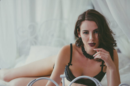 A young attractive girl with dark hair in black lingerie with evening make-up and jewelry and a white bathrobe on a white bed in the bedroom. beauty and fashionの写真素材