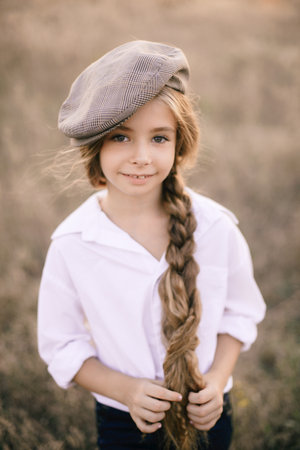 cute little girl with a long braid of blond hair in a white shirt and a men's vintage cap at sunset...
