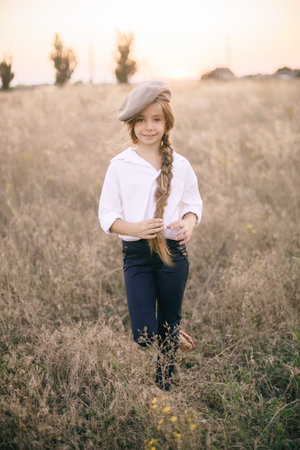 cute little girl with a long braid of blond hair in a white shirt and a men's vintage cap at sunset in a summer field.の写真素材