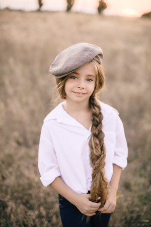 cute little girl with a long braid of blond hair in a white shirt and a men's vintage cap at sunset in a summer field.の写真素材
