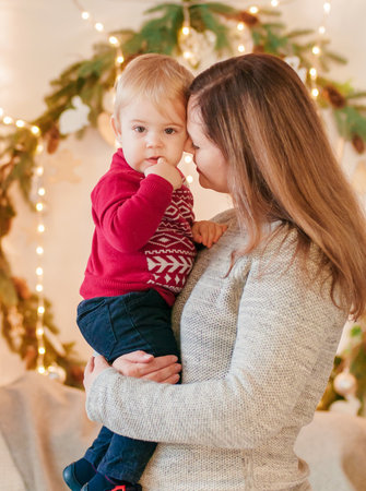A young blond-haired mother with her little son in her arms in a room decorated with pine needles...