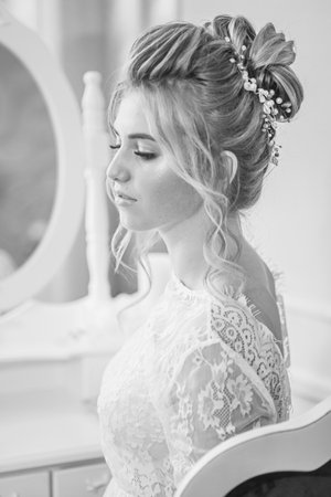 A young beautiful bride with a wedding hairstyle and makeup in the morning in a floor-length white lace tunic near the boudoir table. wedding morningの写真素材