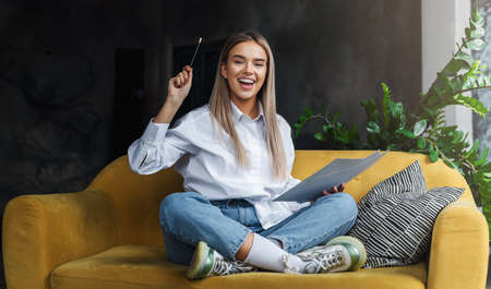 Girl sitting on sofa, drawing on paper with pencil. Cheerful mood in self-isolation, educational,...
