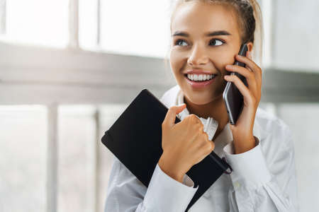 Smiling girl talking on smartphone, holding black notepad. Beautiful girl use mobile phone for work, call clientsの写真素材