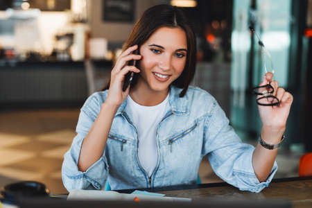 Attractive business woman sits at table in front of laptop and talks on mobile phone, negotiates on the phone.の写真素材