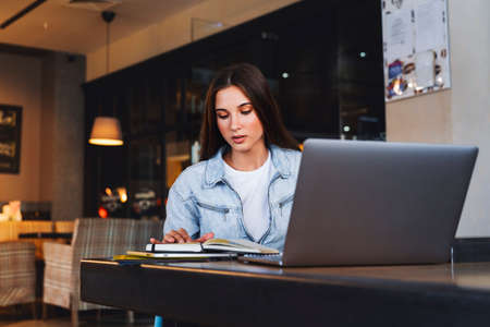 Business woman sits at table in cafe in front of laptop, reads notes from notebook, makes notes in...