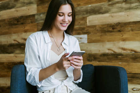 Smiling business woman sitting in armchair against background of wooden wall in loft. Beautiful girl uses mobile phoneの写真素材