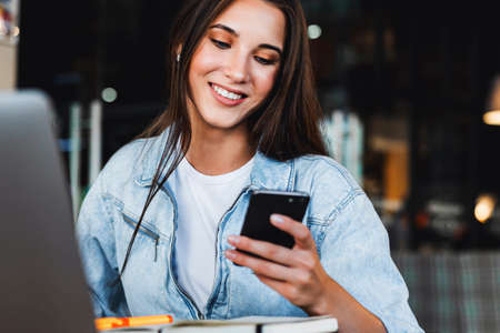 Attractive business woman sits at table in front of laptop and talks on mobile phone, negotiates on...