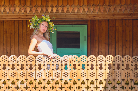 Young beautiful woman in white dress and flower wreath stay in decorative balcony of countryside house at hot and sunny summer dayのeditorial素材