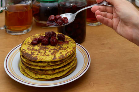 Sweet breakfast: woman hand with spoon pour cherry jam to stack of fresh hot pancakes