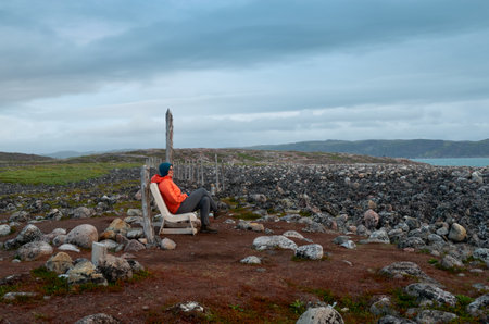 A woman hiker in an orange jacket sits on a plastic chair on a rocky ocean shoreの写真素材