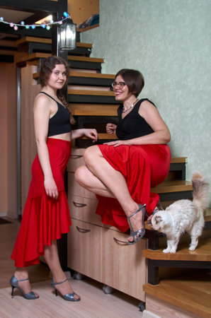 Two female dancers in red skirts and black tank tops are talking in a room next to the stairs and stroking a catの写真素材