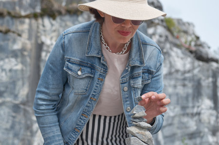 A female tourist in a straw hat and denim jacket builds a zen pyramid of stones against the background of a rockの写真素材