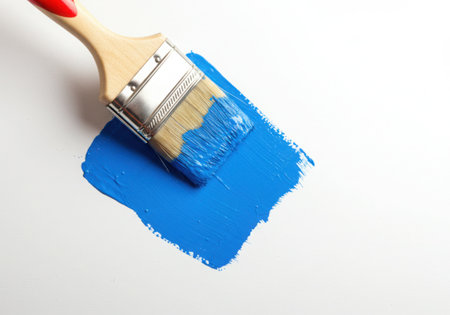 A paint brush with blue paint on its bristles is applying a stroke of blue paint on a white surface. The brush is angled, showing the texture of the bristles and the paint application process.の素材