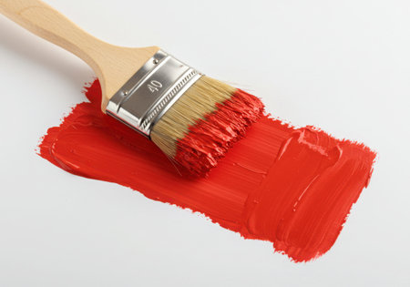 A paint brush with a wooden handle and bristles partially covered in red paint, leaving a vibrant stroke on a white surface. The image captures the essence of painting and creative expression.の素材