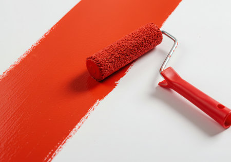 A red paint roller has just painted a stripe of red paint on a white surface. The roller, with its red handle, sits on the edge of the painted area, suggesting a work in progress, perfect for renovation or design concepts.の素材