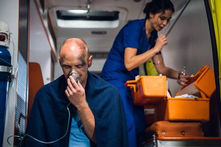 Man breathe through oxygen mask, nurse in uniform opens something taken from medical bag in the ambulance carの写真素材