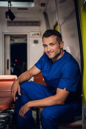 Smiling corpsman in uniform sits and squints in the ambulance car beside stretcherの写真素材