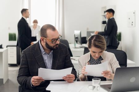 Woman and man in suits sit at a table with documents, other colleagues are standing and talking...