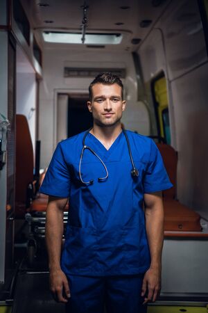 Young doctor stands and looks at the camera in the ambulance car
