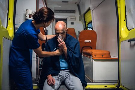 Nurse with stethoscope and man sits in oxygen mask in blanket in the ambulance carの写真素材