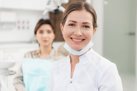 A close up of a female doctor smiling at the camera, with her patient waiting to be checked up in the backgroundの写真素材