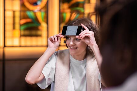 A curly smiling girl with her smartphone on her forehead sitting at the table with friends and...