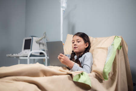 A little girl resting in bed in a hospital ward, she is watching cartoons on her digital tabletの写真素材