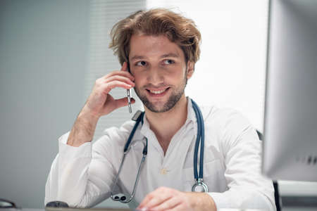 A smiling doctor talking on the phone, while looking at the computer screenの写真素材