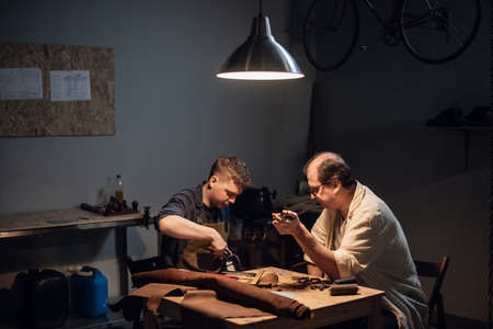 a respectable elderly shoemaker conducts a master class for a young boy on making shoes by hand in a...