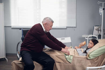 A grandfather is taking care of his grandchild, tucking in her blanket while she is staying in bed...