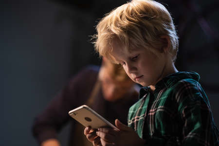 a small blond boy during a break in the labor lesson took a smartphone and communicates on social networksの写真素材
