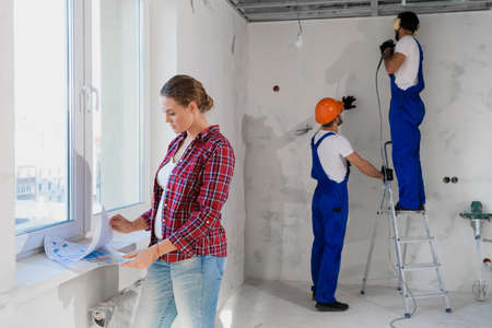 Two workmen in overalls and soundproof headphones drill the wall. The client examines the layout of the apartmentの写真素材