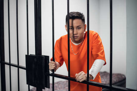 A young asian criminal poses for a photo against the bars in his cell.の写真素材