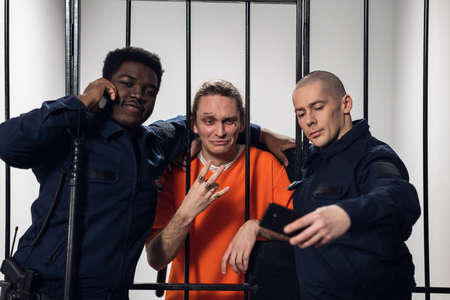Warders in full gear take selfies on a smartphone with a dangerous criminal behind bars. Black guy and white guyの写真素材