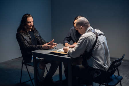 In the police stations interrogation room, good and bad cops are questioning a theft suspect in a leather jacketの写真素材