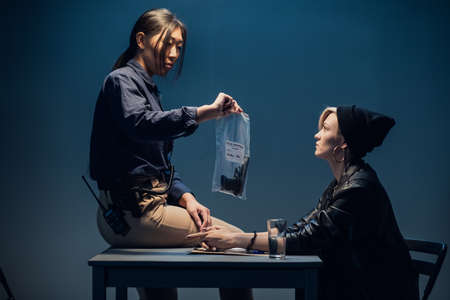 A young asian female investigator sits on a table and shows a package of weapons to a blonde suspect.の写真素材