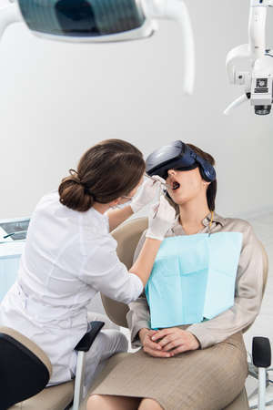 The dentist is doing her job, while the patient is lying with her mouth wide open, wearing VR glasses.の写真素材