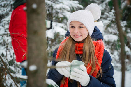 A happy teenage girl enjoys the first snow and drinks hot tea from a mug in the winter forest during the Christmas holidaysの写真素材