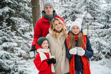 Cozy cute photo of a happy family in winter in a beautiful forest against the background of snowdrifts and snowfall. In their hands they hold a flask and mugs with a hot drinkの写真素材