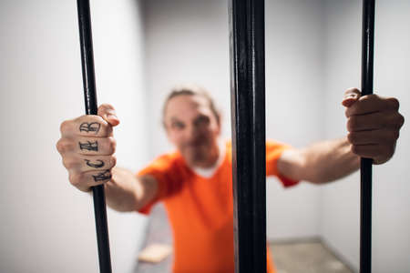 Harsh wild prisoner criminal maniac with tattoos grabs the bars. A close-up portrait in a prison...
