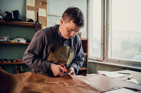 A young tanner in his workshop at the table cuts out leather elements of shoes.