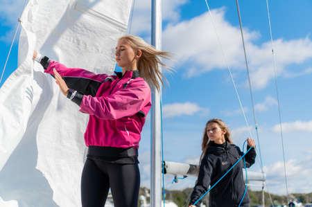 Cute blondes on the pier prepare a sailing yacht for a regatta, pull the sail on the mast with ropes.の写真素材