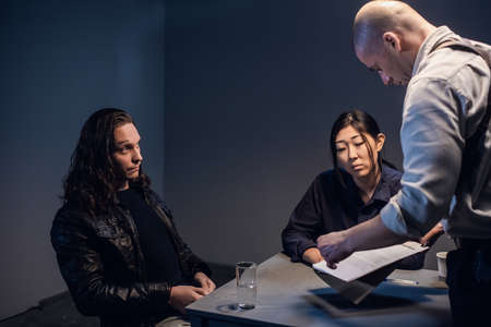 Two police officers are questioning a man with a tattooed face and long hair who is suspected of...