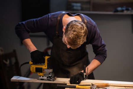 a young carpenter in an apron works in a carpenters shop, making wood products