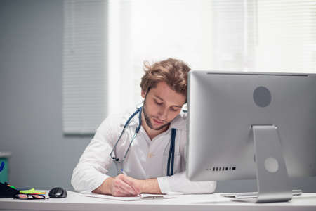 A young male doctor sitting at his desk in the office, working on something.
