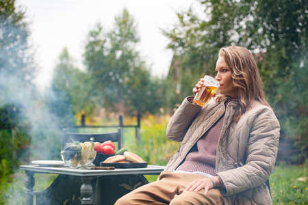 A young blonde woman drinks a light beer from a glass in the backyard of her country house. Cloudy weatherの写真素材