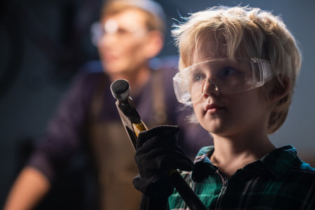 cute blond boy in a carpenters shop in protective glasses and headphones with a hammer, portraitの写真素材