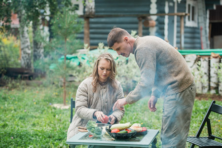 A young couple is preparing for a family barbecue dinner in the courtyard of the house. Cut vegetables on the picnic table. A warm evening in the last days of summerの写真素材